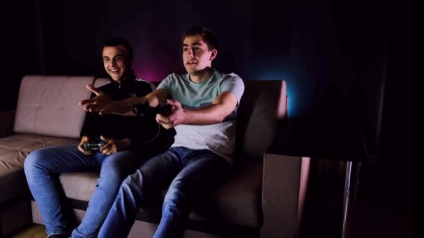 楽しい勝利をお楽しみください 感情的な2人の若い男がテレビの前のソファに座っており ジョイスティックを保持し ゲームコンソールをプレイ興奮しています ワイヤレスコントローラーでメンズゲーム 居心地の良い部屋 — ストック動画