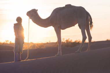 Sahara, Fas, 06-Ocak-2020. Bedevi ve deve kum tepelerinin içinden geçiyor. Sahra Çölü 'nde güneşli güzel bir gün, Fas, Afrika silueti renkli manzaralı göçebe adam..