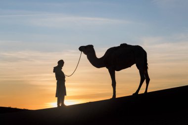Sahara, Fas, 06-Ocak-2020. Bedevi ve deve kum tepelerinin içinden geçiyor. Sahra Çölü 'nde güneşli güzel bir gün, Fas, Afrika silueti renkli manzaralı göçebe adam..