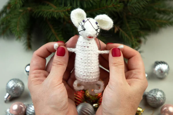Handmade crochet toy. Amigurumi white rat toy in hands on the background of the Christmas tree. Christmas rat is a symbol of the new year 2020.