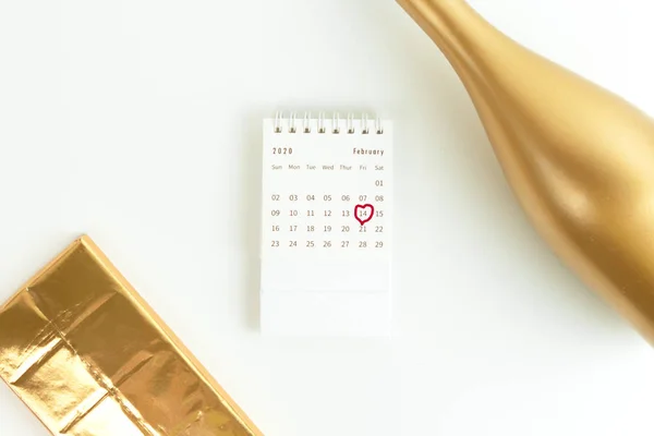 Valentines card, a golden bottle of champagne, chocolate in a gold wrapper and a February calendar with a circled date on a white surface. Top view. — 스톡 사진