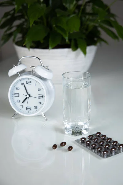White alarm clock and medical pills with a glass of water on the table. Healthcare and medicine. Vitamins and mineral supplements in capsules. Vertical shot.