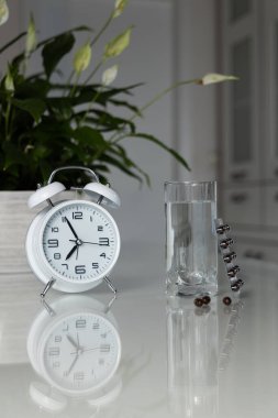 Masanın üzerinde bir bardak su olan beyaz çalar saat ve tıbbi haplar. Sağlık ve ilaç servisi. Vitaminler ve kapsüllerde mineral takviyeleri. Dikey çekim.