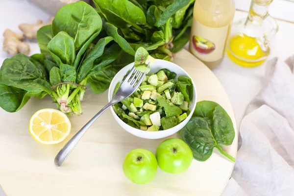 fork with a spinach leaf, and a plate of fresh green vegetables and lettuce leaves is on the table with apples, spinach and vinegar on a light background. Farm organic product, home-made