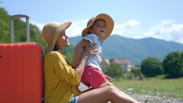 Γυναίκα με παιδί αγόρι σε ένα καπέλο και μια βαλίτσα κάθεται στο δρόμο και περιμένει για ένα αυτοκίνητο στο δρόμο με φόντο τα βουνά και τη φύση. Αναψυχή, Σαββατοκύριακο, οτοστόπ, ταξίδια και τουρισμός έννοια — Αρχείο Βίντεο