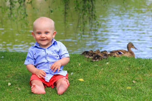 可爱的笑着快乐的小男孩坐在公园的草地上 背后是湖水和鸭子 — 图库照片