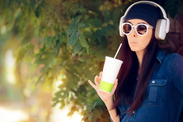 Смішна дівчина в джинсах одягати сонцезахисні окуляри пити сік і слухати музику — стокове фото
