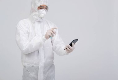 Biyolojik koruyucu üniforma, maske ve eldivenler giyen bilim adamı akıllı telefona virüs bakterilerini dezenfekte etmek için dezenfektan sıkıyor.