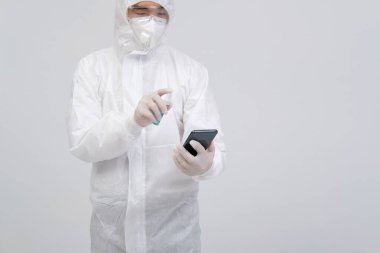 Biyolojik koruyucu üniforma, maske ve eldivenler giyen bilim adamı akıllı telefona virüs bakterilerini dezenfekte etmek için dezenfektan sıkıyor.