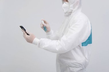 Biyolojik koruyucu üniforma, maske ve eldivenler giyen bilim adamı akıllı telefona virüs bakterilerini dezenfekte etmek için dezenfektan sıkıyor.