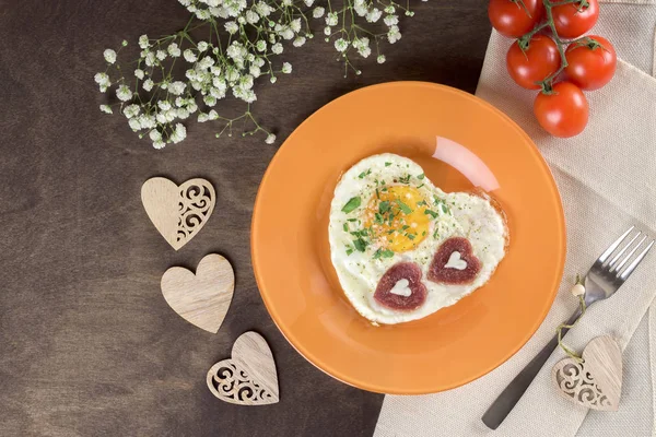 Tasty healthy breakfast for Valentine\'s Day and Mother\'s Day. Fried scrambled eggs heart-shaped on an orange plate with sausages chorizo salami in the shape of a heart. Serving on a wooden table. Homemade food. Top view, horizontal, flatlay