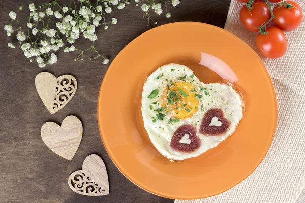 Tasty healthy breakfast for Valentine\'s Day and Mother\'s Day. Fried scrambled eggs heart-shaped on an orange plate with sausages chorizo salami in the shape of a heart with fresh herbs. Serving on a wooden table. Homemade food. Top view, horizontal