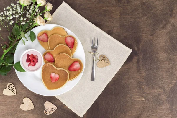 Hausgemachte herzförmige Pfannkuchen mit Erdbeercreme und Beerenscheiben auf einem weißen Teller auf einem Holztisch. Frühstück im rustikalen Stil oder Brunch zum Valentinstag. Ansicht von oben, Kopierraum — Stockfoto