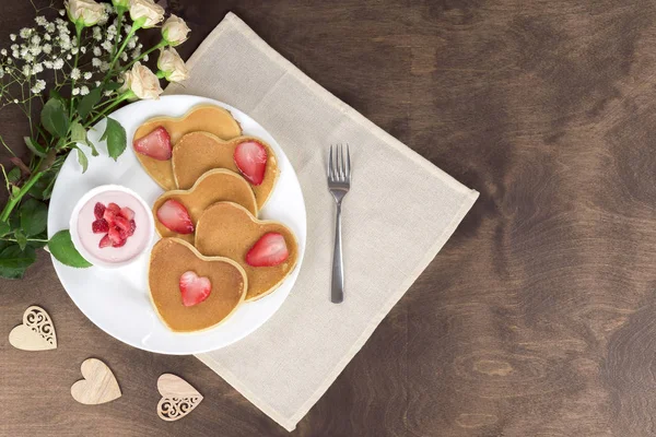 Homemade heart shaped pancakes with strawberry cream and berry slices on a white plate served with a dessert fork on a wooden table. Rustic style breakfast or brunch. Top view, copyspace — Stock Photo, Image