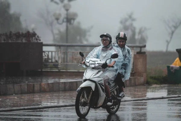 Двое азиатских мужчин едут на скутере под проливным дождем в маленьком горном городке Сапа, Северный Вьетнам, прикрываясь, чтобы защитить себя — стоковое фото