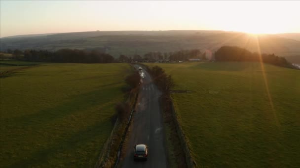 Съемки с воздуха, следующие за автомобилями, едущими по узкой сельской дороге через сельскохозяйственные угодья в Национальном парке Пик-Дистрикт, Великобритания, во время красивого заката, Зима 2019 — стоковое видео