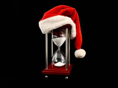 Hourglass in the cap of Santa Claus. Christmas hat on glass hourglass. Glass device for measuring time, on a black background. Concept: outgoing year