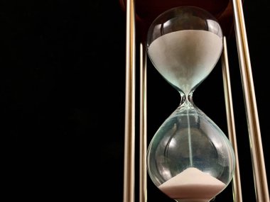 Hourglass on a stand, black background. Glass hourglass in the case. Glass time meter. Concept: time is running out, time management