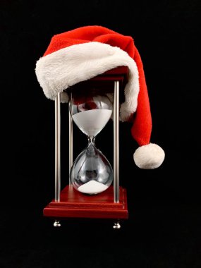 Hourglass in the cap of Santa Claus. Christmas hat on glass hourglass. Glass device for measuring time, on a black background. Concept: outgoing year