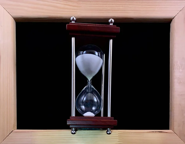 Hourglass in a wooden frame, on a black background. Glass hourglass in a frame for a picture. Glass time meter. Concept: time is running out, time management — Stock Photo, Image