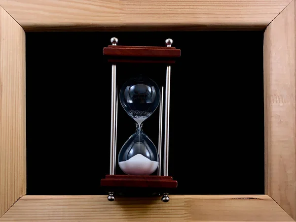 Пісочний годинник в дерев'яній рамці, на чорному тлі. Скляний пісочний годинник у рамці для картини. Вимірювач часу скла. Концепція: час закінчується, управління часом — стокове фото