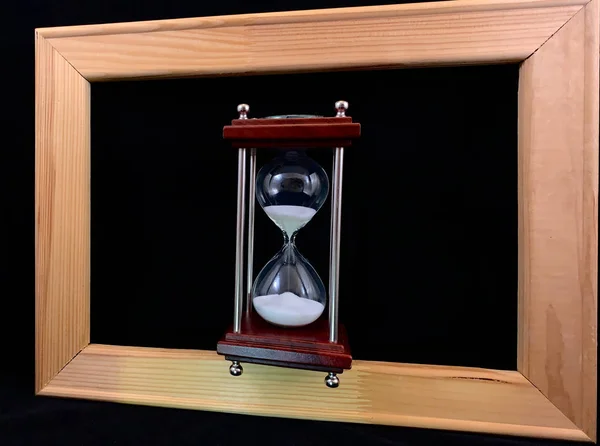 Hourglass in a wooden frame, on a black background. Glass hourglass in a frame for a picture. Glass time meter. Concept: time is running out, time management — Stock Photo, Image