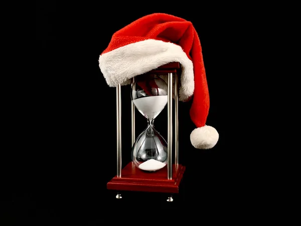 Hourglass in the cap of Santa Claus. Christmas hat on glass hourglass. Glass device for measuring time, on a black background. Concept: outgoing year — стокове фото