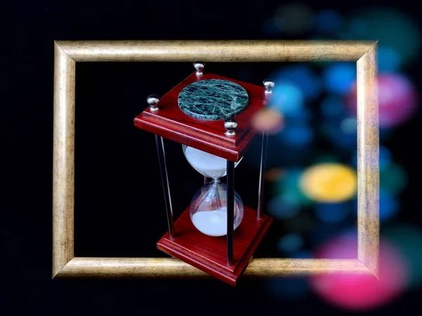 Hourglass in a wooden frame, on a black background. Glass hourglass in a frame for a picture. Glass time meter. Concept: time is running out, time management