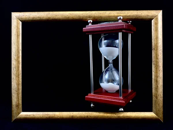 Hourglass in a wooden frame, on a black background. Glass hourglass in a frame for a picture. Glass time meter. Concept: time is running out, time management — 图库照片