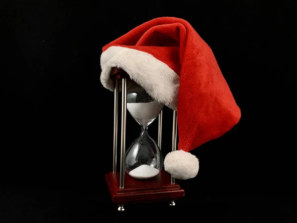 Hourglass in the cap of Santa Claus. Christmas hat on glass hourglass. Glass device for measuring time, on a black background. Concept: outgoing year — Stock Photo, Image