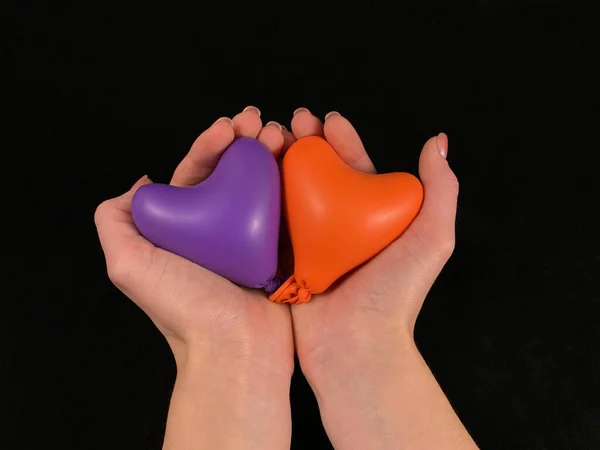 Heldere opblaasbare ballonnen in de vorm van een hart in de handen. Twee harten in vrouwelijke handpalmen, op een zwarte achtergrond. Meisje houdt een rubberen hart. — Stockfoto