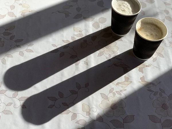 Cangkir kertas sekali pakai dengan cappuccino dan bayangan di atas meja. Sebuah porsi kopi hitam dengan susu untuk dua . — Stok Foto