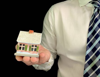 Bir adam elinde oyuncak bir ev tutuyor. Beyaz gömlekli, siyah arka planda evin modelini taşıyan bir adam. Konsept: emlak yatırımı ve ipotek, konut inşaatı, satın alma