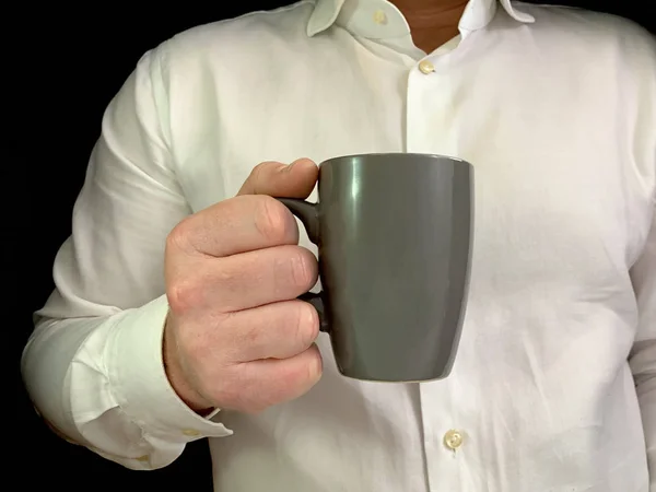 Un hombre tiene una taza de café gris en sus manos. Un hombre con una camisa blanca con una taza de té en la mano, sobre un fondo negro. Concepto: tiempo para tomar café, descanso . — Foto de Stock