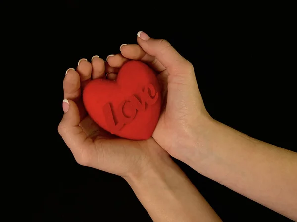 L'iscrizione "Love" e un cuore rosso nelle mani di una ragazza su uno sfondo nero. Una donna tiene un segno cardiaco con le dita. Concetto: San Valentino, matrimonio e altre festività . — Foto Stock