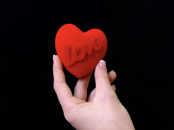 L'iscrizione "Love" e un cuore rosso nelle mani di una ragazza su uno sfondo nero. Una donna tiene un segno cardiaco con le dita. Concetto: San Valentino, matrimonio e altre festività . — Foto Stock