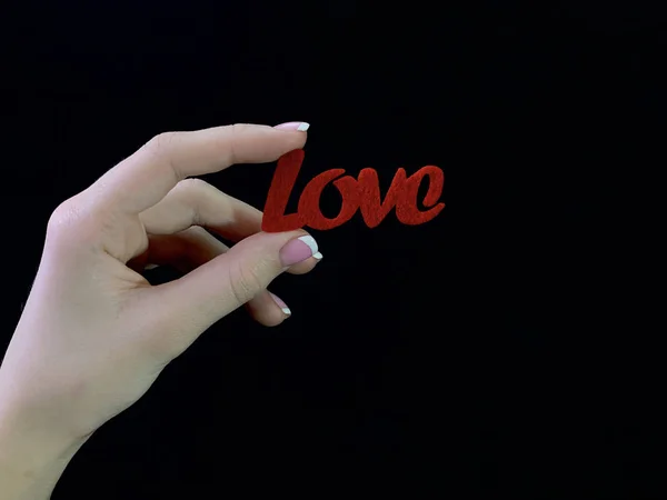 La inscripción "Amor" y un corazón rojo en las manos de una niña sobre un fondo negro. Una mujer sostiene un signo del corazón con los dedos. Concepto: Día de San Valentín, boda y otros días festivos . — Foto de Stock