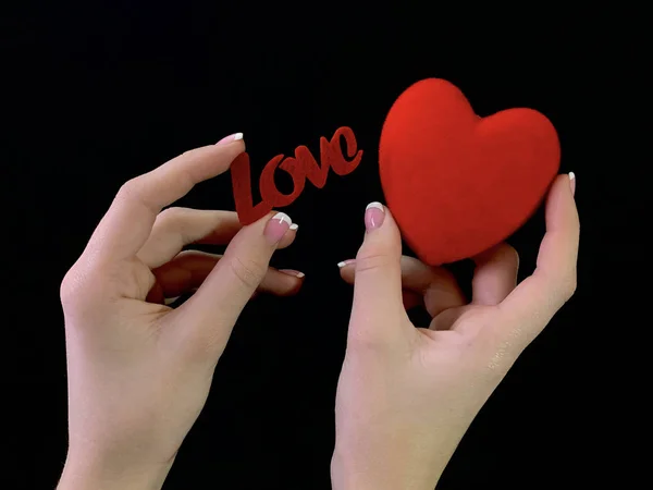 A inscrição "Amor" e um coração vermelho nas mãos de uma menina em um fundo preto. Uma mulher está segurando um sinal de coração com os dedos. Conceito: Dia dos Namorados, casamento e outros feriados . — Fotografia de Stock