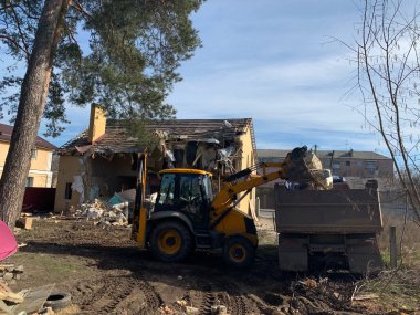 Yıkılmış tuğla evin yanında ağır ekipman var. Evin harabelerinin arka planında inşaat makineleri var. Kazıcılı eski bir evin yıkımı. Binanın ve kamyonun yıkılmış duvarları..