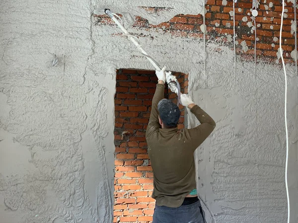 The craftsman puts the plaster on a brick wall. The builder is engaged in wall cladding. The painter - the plasterer works with a spray gun.