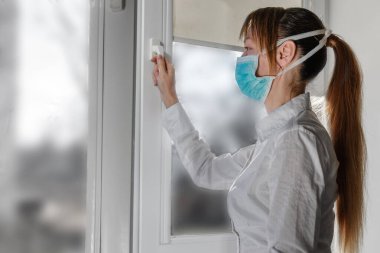 Tıbbi maskeli bir kadın hava almak için bir pencere açar. COVID-19 koronavirüs salgınını önlemek için kendini izole etme.