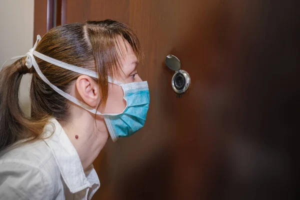 a female in medical green mask looks through the peephole. Self-isolation to prevent the coronavirus pandemic.
