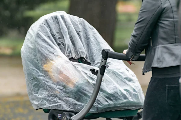 Bir anne bebeğiyle birlikte yağmurlu bir bahar gününde bir şehir parkında film şeridine sarılı bir şekilde yürür..