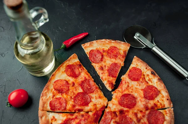 Sliced pizza with pizza cutter, vegetables and olive oil on black stone background.
