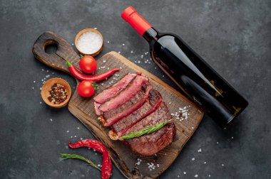 Bir şişe şarap, ızgara biftek, baharatlar, domatesler ve siyah arka planda otlar.