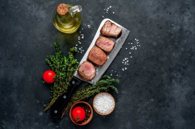 ızgara madalyon biftek dilimleri, otlar, zeytinyağı ve gri kırsal masa arka planında biberiye dalları.