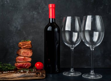 Biftek madalyonu, otlu dilimler ve gri kırsal masa arka planında bir şişe kırmızı şarap.