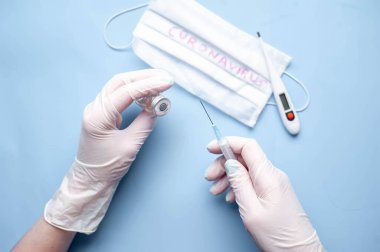 Doktor ellerinde aşı ve koronavirüsten şırınga bulunan tıbbi eldivenleri mavi arka planda tutuyor.