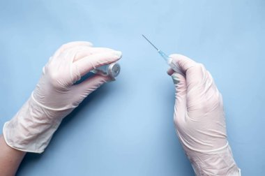 Doktor ellerinde aşı ve koronavirüsten şırınga bulunan tıbbi eldivenleri mavi arka planda tutuyor.