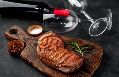 Kesme tahtasında baharatlı ızgara biftek ve bir şişe kırmızı şarap, arka planda bardaklar.